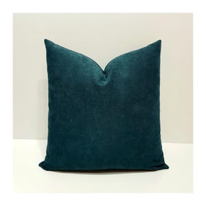 May include: A square throw pillow featuring a solid deep teal color with a soft, textured fabric finish. The pillow has a distinct, plump shape with a slight indentation at the top center, creating a clean and uniform appearance for home decor.