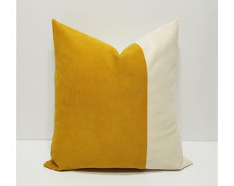 May include: A square throw pillow featuring a color-blocked design with a large mustard yellow section on the left and a smaller cream-colored section on the right. The fabric has a soft, matte texture and the pillow is filled to a plump shape.