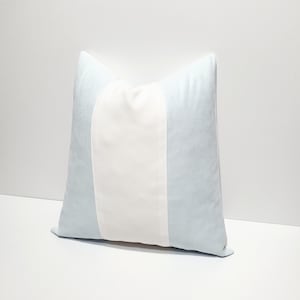 May include: A square throw pillow featuring a vertical color-blocked design. The pillow has a wide, solid white center panel flanked by two light blue side panels. The fabric appears to have a soft, smooth texture throughout the entire surface.