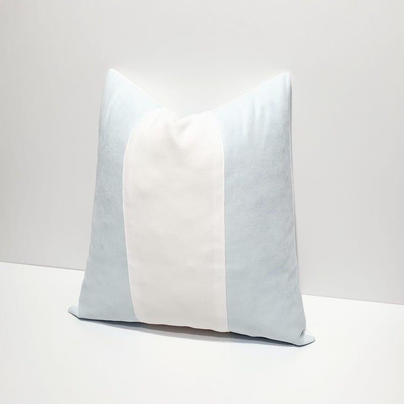 May include: A square throw pillow featuring a vertical color-blocked design. The pillow has a wide, solid white center panel flanked by two light blue side panels. The fabric appears to have a soft, smooth texture throughout the entire surface.