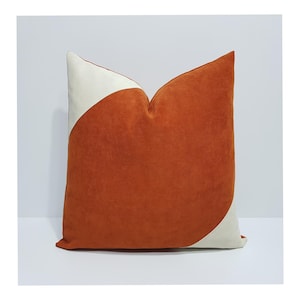 Puede incluir: Un cojín cuadrado con un diseño de dos tonos que presenta una gran sección curva de tela color naranja quemado contrastada por tela color blanco hueso en las esquinas superior izquierda e inferior derecha. El material tiene una textura mate.
