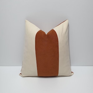 May include: A square throw pillow featuring a color-blocked design with a wide, vertical, textured rust-orange center panel flanked by two smooth, cream-colored side panels. The pillow has a distinct, pointed top edge and a clean, structured appearance.