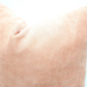 May include: A square throw pillow cover in a solid, muted peach-pink color. The fabric features a fine, uniform woven texture with a soft, matte appearance, showing a subtle depth in the light and shadow across the surface of the pillow.