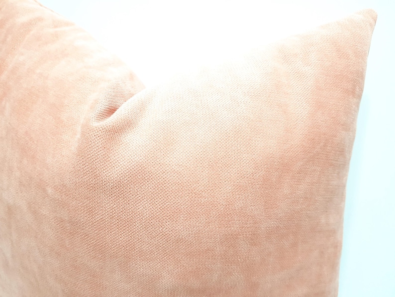 May include: A square throw pillow cover in a solid, muted peach-pink color. The fabric features a fine, uniform woven texture with a soft, matte appearance, showing a subtle depth in the light and shadow across the surface of the pillow.