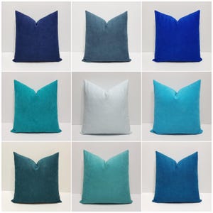 May include: A collection of nine square throw pillows in various shades of blue, including navy, royal blue, teal, turquoise, and light gray. Each pillow features a soft, solid-colored fabric with a distinct pinched top center detail.