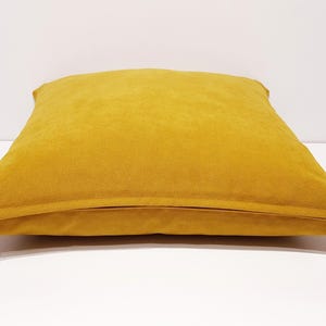 May include: A square mustard yellow throw pillow with a smooth, solid-colored fabric cover. The pillow features a visible horizontal seam along the bottom edge where a white zipper pull is attached, allowing for the removal of the cover.