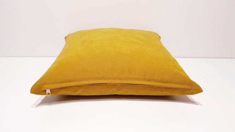 May include: A square mustard yellow throw pillow with a smooth, solid-colored fabric cover. The pillow features a visible horizontal seam along the bottom edge where a white zipper pull is attached, allowing for the removal of the cover.