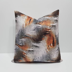 May include: A decorative cushion with an abstract pattern in shades of brown, orange, and grey. The cushion has a textured fabric and a square shape.