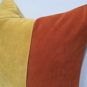 May include: A square throw pillow featuring a color-blocked design with a vertical seam. The left side is a mustard yellow fabric with a fine, textured weave, while the right side is a solid burnt orange fabric with a similar textured finish.
