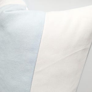 May include: A square throw pillow featuring a color-blocked design with a vertical panel of light blue textured fabric on the left side and a larger panel of solid white fabric on the right side, showcasing a clean and simple aesthetic.