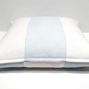 May include: A square throw pillow featuring a vertical color-blocked design. The pillow has a wide, light blue central panel flanked by two white side panels. A visible zipper closure is located along the bottom edge of the pillow cover.