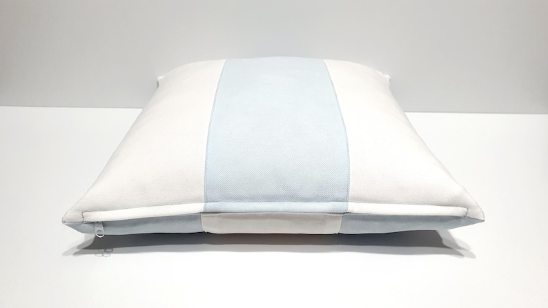 May include: A square throw pillow featuring a vertical color-blocked design. The pillow has a wide, light blue central panel flanked by two white side panels. A visible zipper closure is located along the bottom edge of the pillow cover.