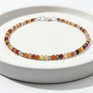 May include: A delicate beaded bracelet featuring small, faceted, multi-colored gemstones in shades of orange, amber, brown, and cream, interspersed with tiny, polished silver-toned metal beads. The bracelet is secured with a silver-toned metal clasp.