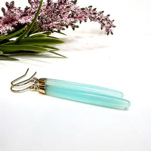 Peut inclure: Une paire de boucles d'oreilles allongées bleu clair avec des accents dorés. Les boucles d'oreilles sont présentées sur une surface blanche, avec une branche de fleurs violettes et vertes en arrière-plan. Design épuré et minimaliste.