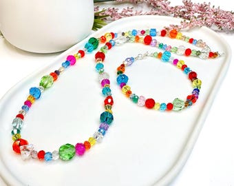 Op de afbeelding: Een kleurrijke kralenketting en armband set. De ketting en armband hebben een verscheidenheid aan levendige kralen in rood, groen, blauw, geel en roze. De sieraden worden gepresenteerd op een wit dienblad.