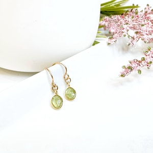 May include: A pair of gold earrings with small, round, light green gemstones. The earrings have a simple, delicate design.