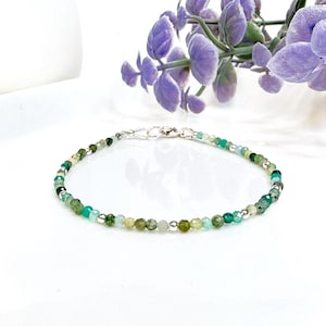 May include: A delicate bracelet made with green and blue gemstone beads and silver accents. The bracelet is on a white surface with a purple flower in the background.
