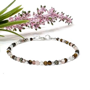 May include: Delicate beaded bracelet featuring a variety of small, faceted gemstones in shades of brown, pink, clear, and gray. The bracelet is strung on a silver chain and has a silver clasp. The background includes pink and green floral accents.