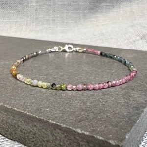 May include: A delicate bracelet made with small, colorful gemstones. The bracelet features a gradient of colors, starting with light green and yellow beads, transitioning to dark green and black beads, and ending with pink beads. The bracelet is secured with a silver clasp.