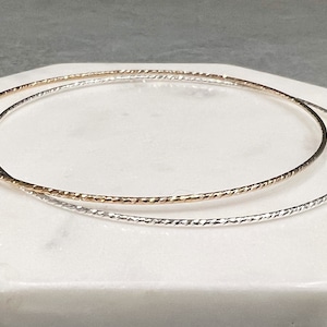 May include: Two thin bangle bracelets, one silver and one gold, with a textured, braided design.