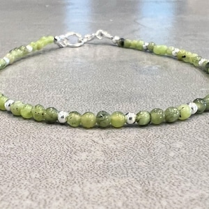 May include: A delicate bracelet featuring small, round, light green beads interspersed with silver-colored beads. The bracelet is secured with a silver-colored clasp. The beads have a speckled appearance.