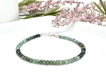 May include: A delicate bracelet featuring small, faceted emerald beads in shades of green and gray. The beads are strung together with a silver clasp. The bracelet is displayed on a white surface with floral accents.