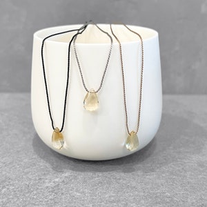 May include: Three necklaces with teardrop-shaped, light yellow gemstones. Each pendant hangs from a different colored cord: black, silver, and gold. The necklaces are displayed against a white ceramic background.