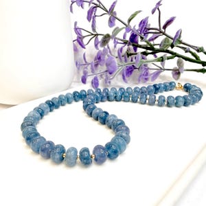 Pode incluir: Um colar composto por contas redondas de pedra preciosa azul polida dispostas numa única fila. Pequenas contas espaçadoras metálicas estão posicionadas entre cada uma das pedras azuis maiores. O colar apresenta um fecho de metal dourado.
