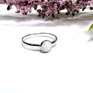 May include: A silver ring featuring a single, round, bezel-set opal cabochon. The band is smooth and polished, showcasing a metallic luster. The opal displays iridescent flashes of color, including white, blue, and green tones, set within a simple, circular metal frame.