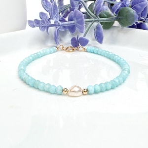 May include: A delicate bracelet made with light blue gemstone beads and a single white pearl accent. The bracelet is finished with a gold clasp.