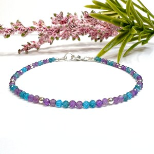 May include: A delicate bracelet featuring alternating faceted beads in shades of purple and turquoise, interspersed with small silver-colored beads. The bracelet is secured with a silver clasp. The background includes a sprig of lavender and greenery.