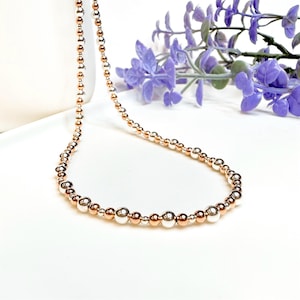 May include: A delicate necklace featuring alternating silver and rose gold beads. The necklace is displayed on a white surface with a sprig of purple flowers in the background. The beads vary in size, creating a subtle, elegant design.