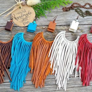 May include: A selection of handmade leather fringe earrings in shades of brown, turquoise, orange, white, and red. Each earring has a copper hoop and a leather detail at the top. A tag with the words "handmade with love" is visible.