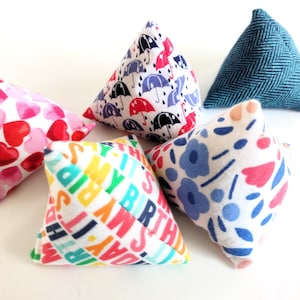 May include: Five small, triangular fabric items. One has a blue herringbone pattern, another features a colourful "Happy Birthday" print. The others display patterns of hearts, umbrellas, and flowers in various colours.