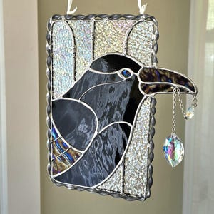 May include: Stained glass raven art piece with a black and iridescent body, a dark brown beak, and a sparkling crystal charm hanging from its beak. The piece is framed with a silver border and has a white ribbon for hanging.