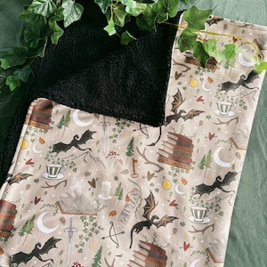 May include: A beige blanket with a black, fluffy reverse. The patterned side displays dragons, books, swords, teacups, and the text "Enemies-Lovers" and "Batty Books". Green ivy decorates the blanket's edges.