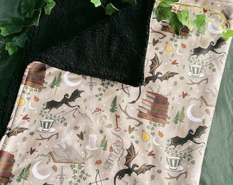 Può includere: Una coperta beige con un rovescio nero e soffice. Il lato superiore presenta un motivo ripetuto di draghi, libri, spade, tazze da tè e i testi "Enemies-Lovers" e "Batty Books". Decorata con edera verde.