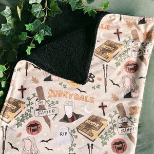 May include: A beige and black blanket featuring a repeating pattern of crosses, bats, books, and character illustrations. The blanket has the words "Sunnydale" and "The Slayer" printed on it. The reverse side is black.