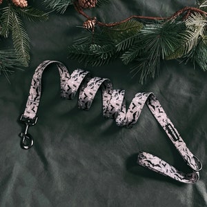 May include: A grey and black patterned dog lead with a black metal clip. The lead has a repeating pattern of bats and crescent moons.