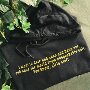 May include: Black hooded sweatshirt with a yellow graphic print that reads "I want to date and shop and hang out, and save the world from unspeakable evil. You know, girly stuff."