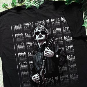 May include: A black t-shirt featuring a graphic of a person with sunglasses playing an electric guitar. The background includes the repeated white text "i think you're scared of being basic" in a gothic font, layered behind the central figure.