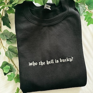 May include: Black crewneck sweatshirt with the embroidered text "who the hell is bucky?" in white. The sweatshirt is folded and placed on a white surface with green foliage in the background.