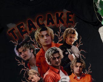 May include: Black t-shirt featuring a collage of images of a person with blonde hair, with the word "TEACAKE" in red dripping letters. The design includes lightning bolt graphics and images of the person in various poses.