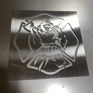 May include: A metal stencil of a firefighter in a helmet and coat, holding an axe, standing in front of a fire department cross. The stencil is cut out of a silver metal sheet.