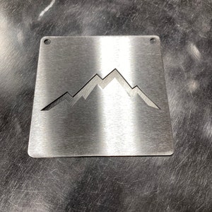 May include: A silver metal square sign with a cut-out mountain range design. The sign has two holes at the top for hanging.
