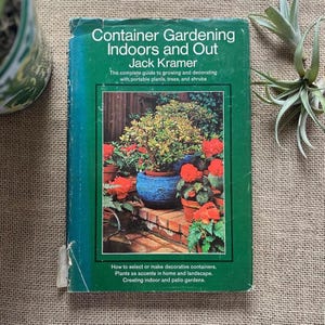 May include: A well-worn book titled "Container Gardening Indoors and Out" by Jack Kramer. The cover features a vibrant image of potted plants and flowers. The book's subtitle promises guidance on growing and decorating with portable plants, trees, and shrubs.