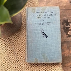 May include: A vintage field guide titled "A Field Guide to the Birds of Britain and Europe." The book has a light blue cover with the title and author's names in dark blue. A black and white bird illustration is also on the cover. The book is resting on a burlap surface.