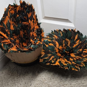 Peut inclure: Deux pièces décoratives en plumes. L'une est en forme de demi-sphère, l'autre est un cercle plat. Les deux sont faites de plumes orange, vertes et noires fixées sur une base en carton. Les pièces sont sur un tapis brun clair.