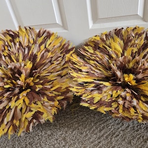 Peut inclure: Deux chapeaux Juju décoratifs. Chaque chapeau est rond et fait de plumes dans les tons de jaune, marron et crème. Les chapeaux sont exposés sur un tapis de couleur neutre devant une porte blanche.