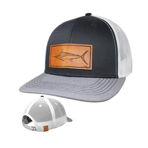 May include: A baseball cap featuring a black front panel, a grey brim, and white mesh back panels. A rectangular tan leather patch is centred on the front, displaying a detailed engraved illustration of a marlin fish. A snapback closure is visible.